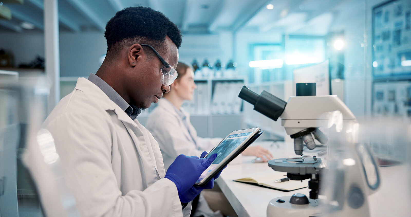 A researcher in a lab coat reviews data on a tablet beside a microscope.